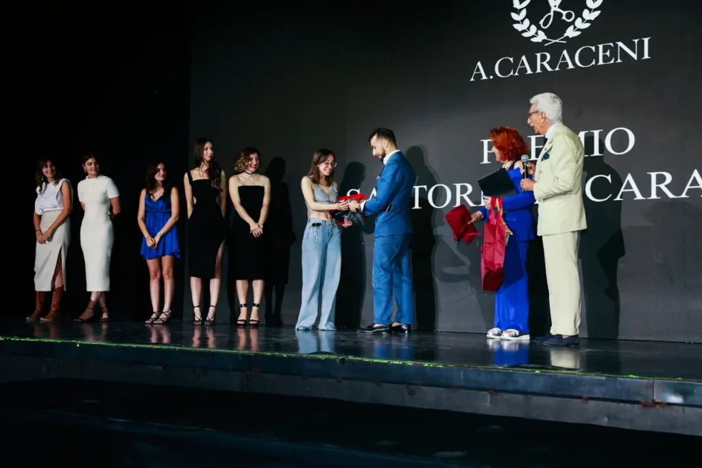 studenti su un palco scuro con un muro scuro che ricevono una targa con premio A.Caraceni