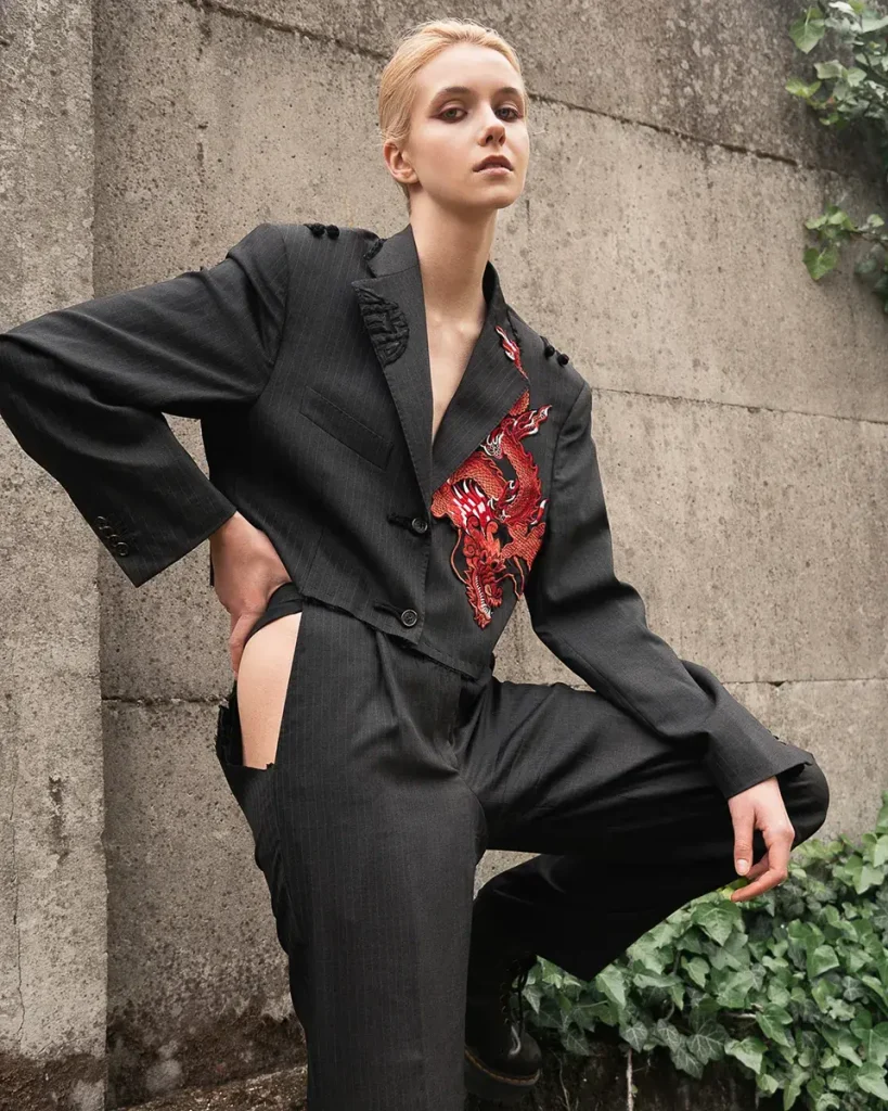 Modella con un completo nero posa con dietro un muro.