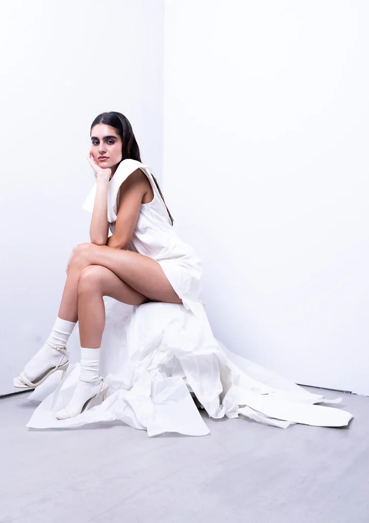 Modella in posa seduta in studio indossa un abito bianco con voluminosi drappeggi.