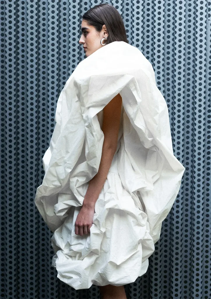 Modella in posa in ambiente industriale indossa un abito bianco con voluminosi drappeggi.